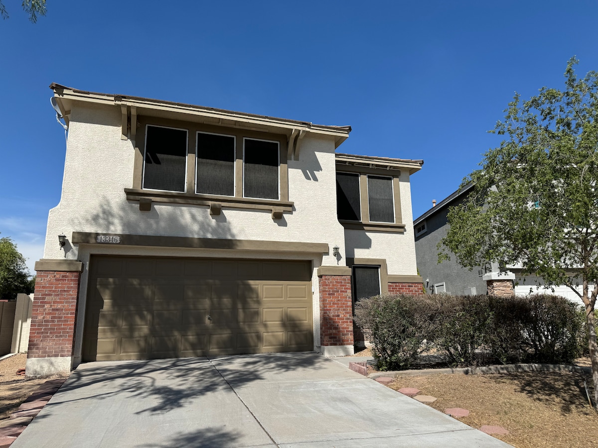 The front exterior of the home is shown, featuring a two-story structure with a beige facade and dark brown accents. Large windows are highlighted on the upper level, and a two-car garage is visible. A landscaped yard with low shrubs and a clear blue sky completes the scene.
