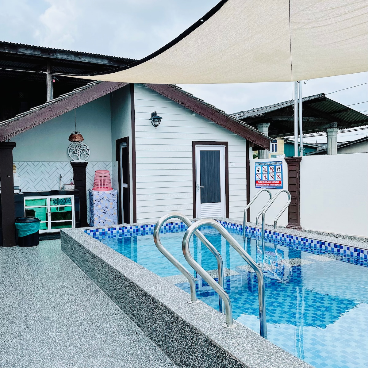 A private pool is bordered by a textured surface, featuring steps leading into the water. Attached to the pool area, a small kitchenette is visible with aqua-colored cabinetry. A shaded lounging area is positioned nearby, providing comfort and relaxation.