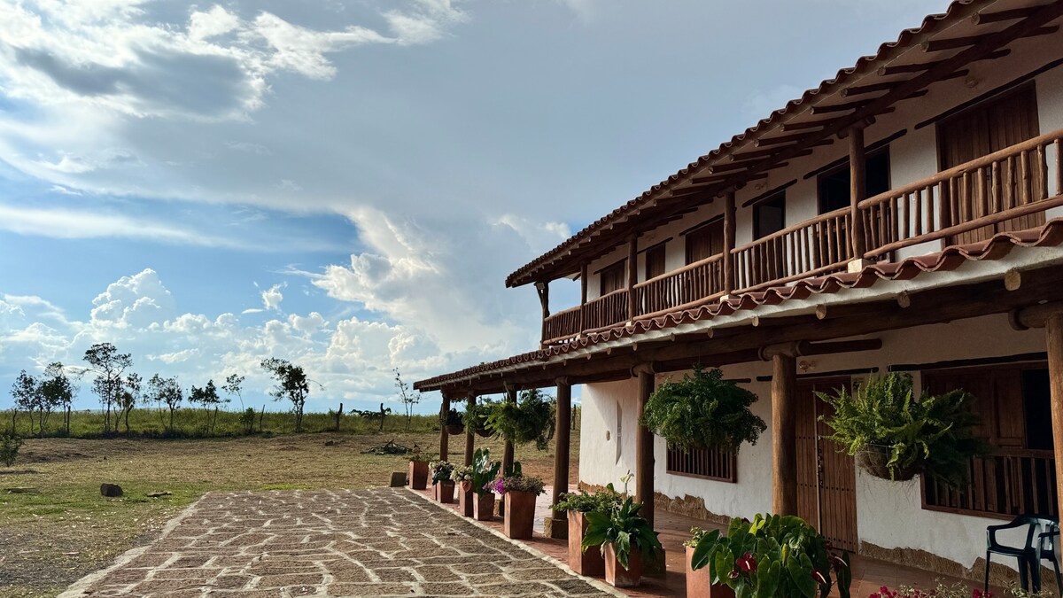 A rustic cabin exterior is presented, featuring wooden balconies and a spacious stone path leading to the entrance. Potted plants adorn the façade, while a serene landscape with trees and open skies is visible in the background.