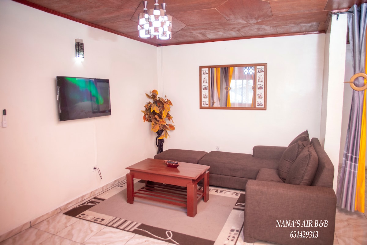 A comfortable living area is shown, featuring a dark sectional sofa positioned next to a wooden coffee table. A flat-screen television is mounted on the wall, while a decorative plant adds a touch of greenery. Large windows are adorned with vibrant curtains that allow natural light.