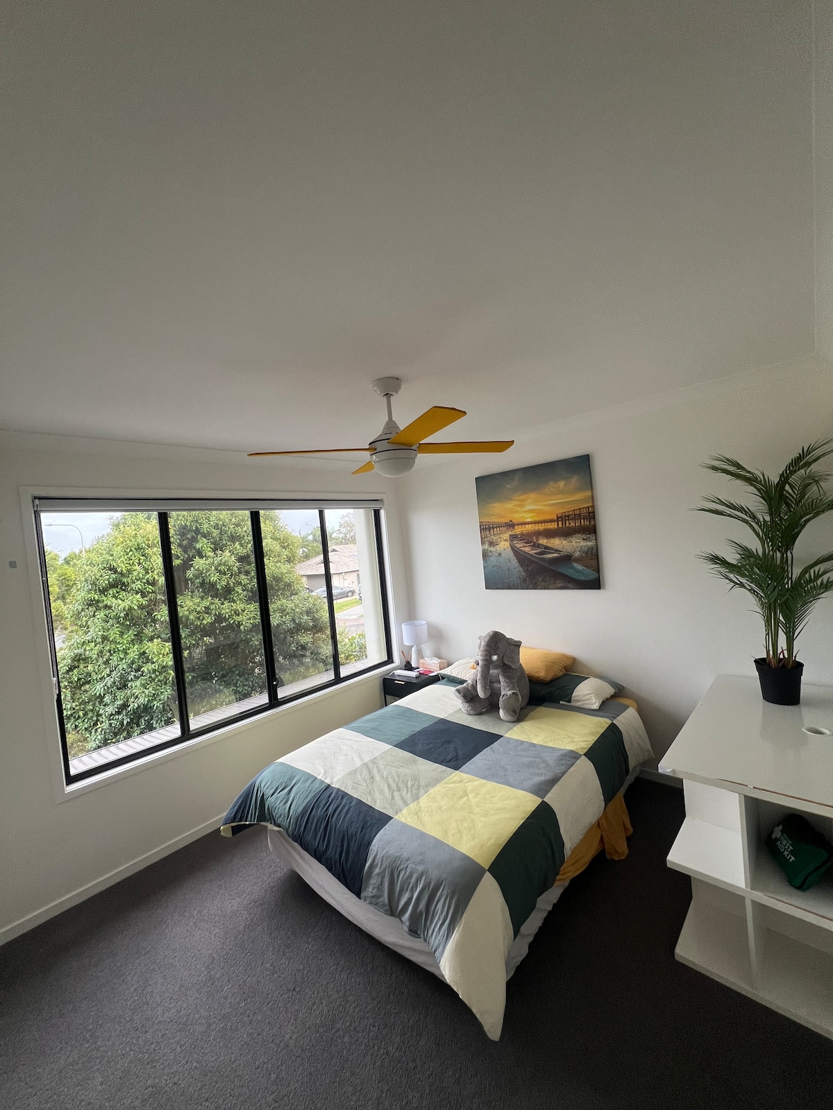 A cozy bedroom features a spacious bed with a patterned quilt and decorative pillows. A ceiling fan is positioned above, and natural light streams through a large window, offering a view of greenery outside. A small nightstand and a potted plant complete the room.