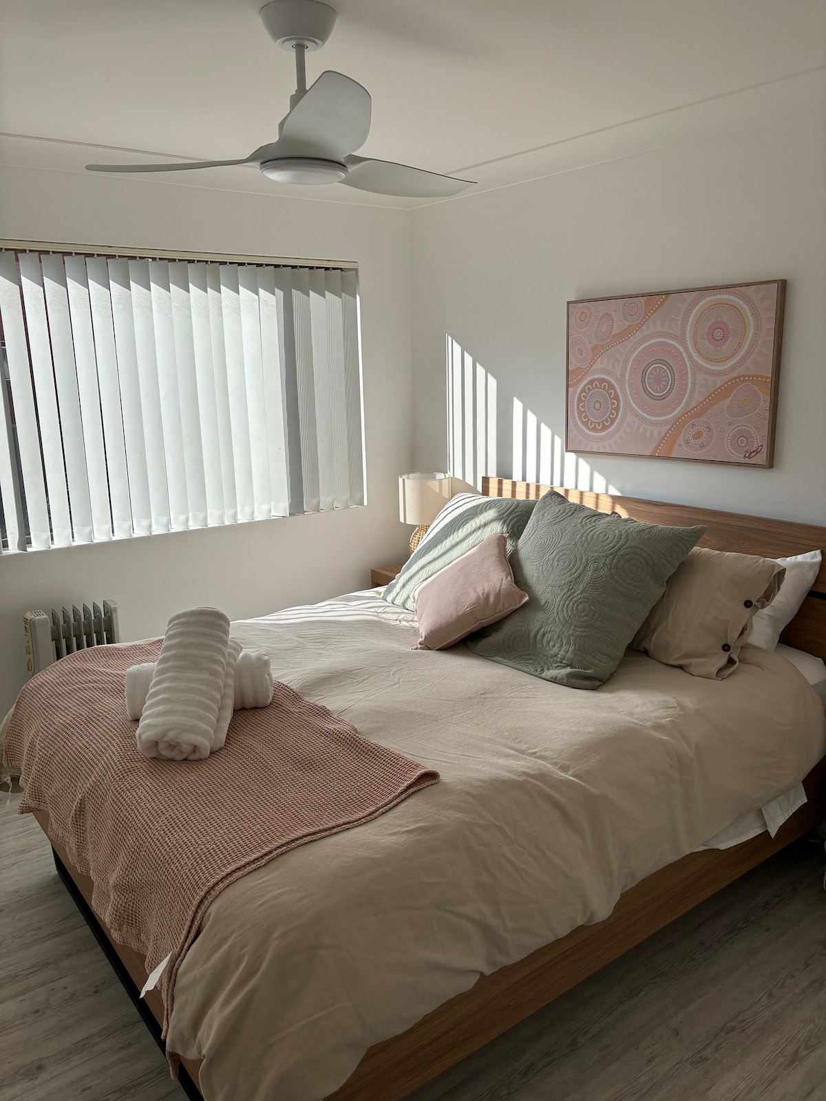 A well-lit bedroom features a spacious bed dressed in neutral tones with decorative pillows and a cozy blanket. Natural light filters through vertical blinds, creating soft shadows. A piece of wall art adds a touch of color, while a ceiling fan enhances the room's comfort.