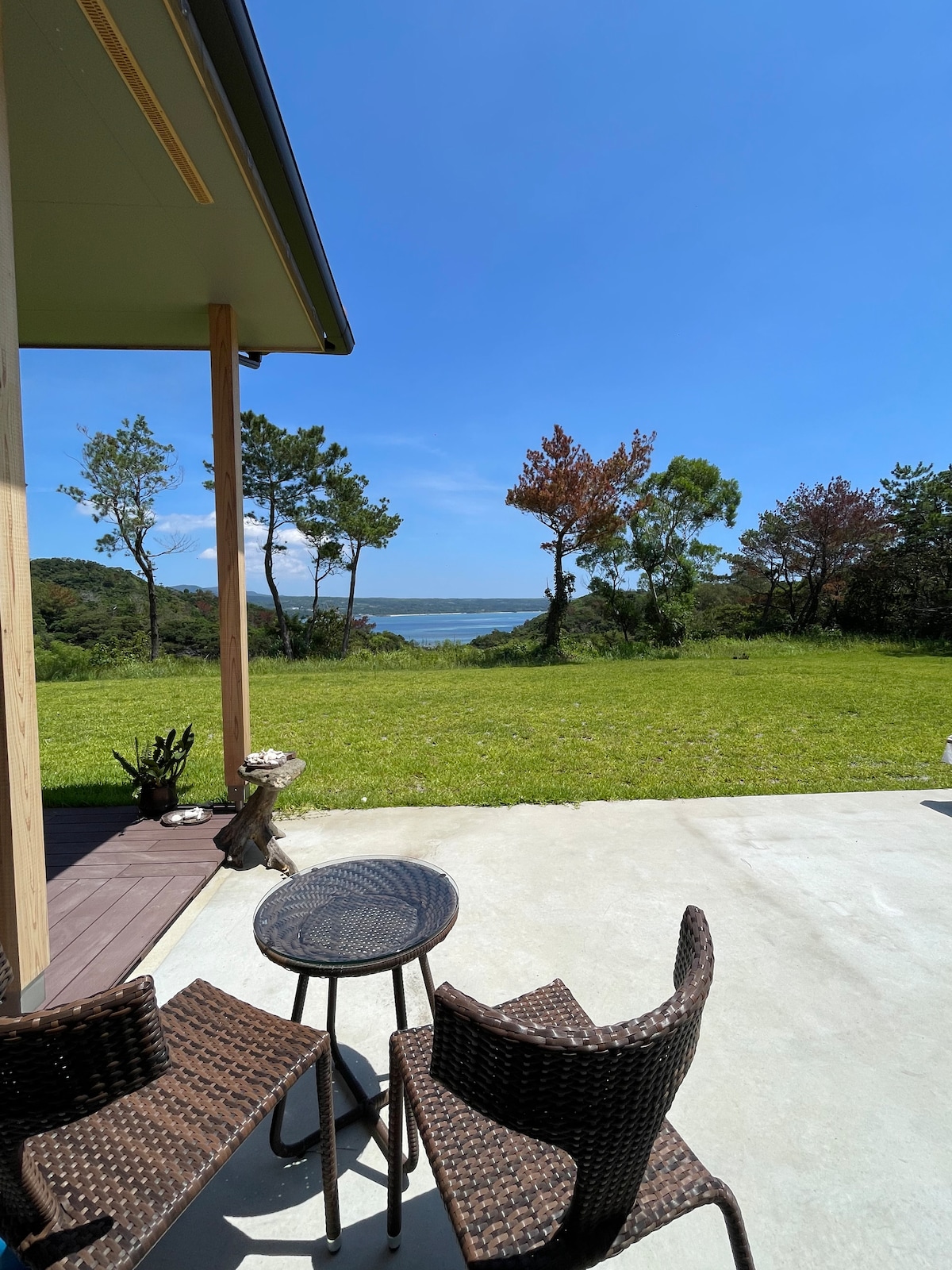 A private outdoor seating area is set on a concrete patio, featuring two wicker chairs and a small round table. The expansive view showcases lush greenery and a serene landscape, with a glimpse of water in the background under a clear blue sky.