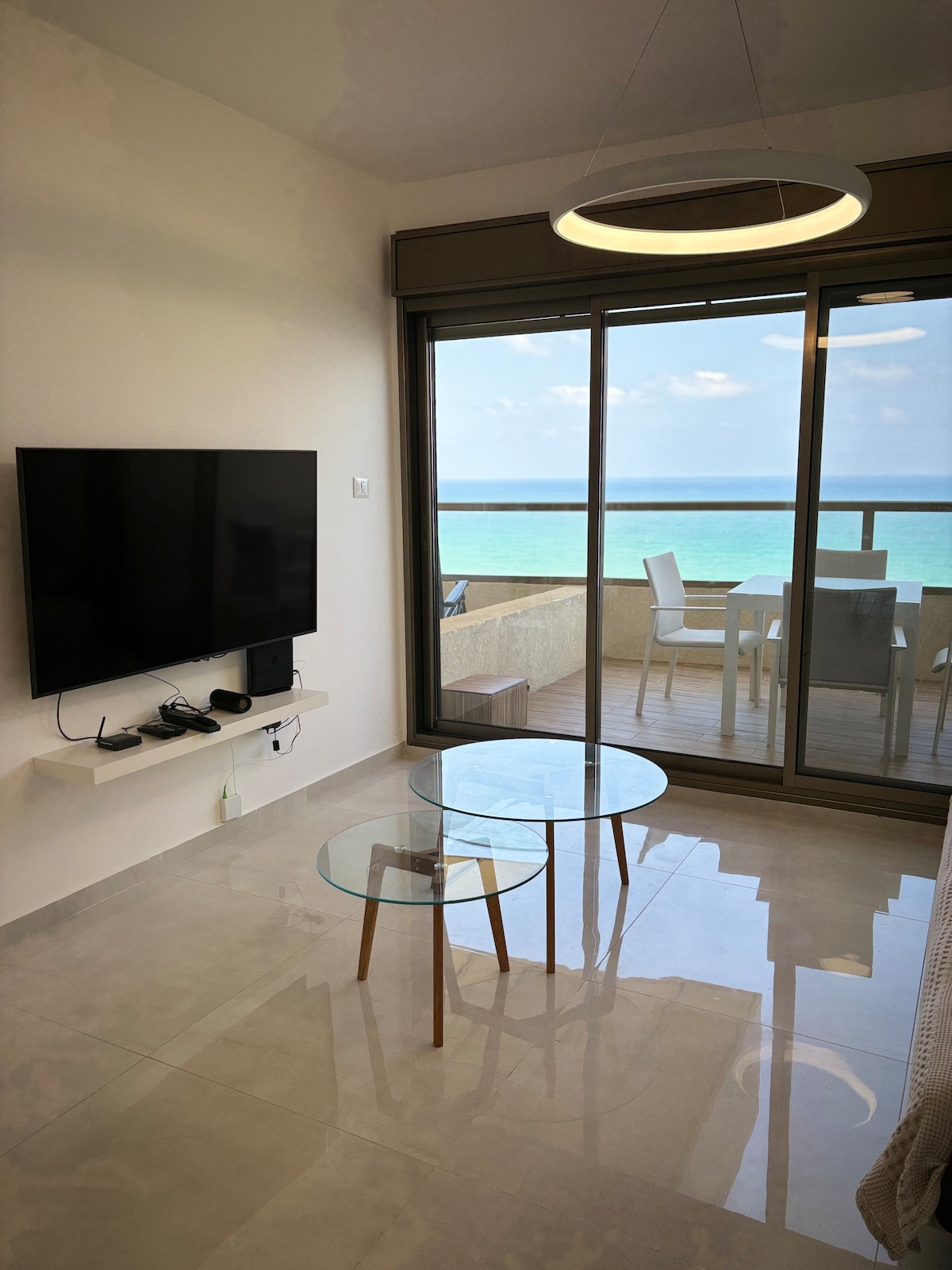 A living area is showcased with a wall-mounted television and two glass coffee tables. Floor-to-ceiling windows provide a stunning view of the ocean. A balcony is visible in the background, featuring outdoor seating for relaxation.