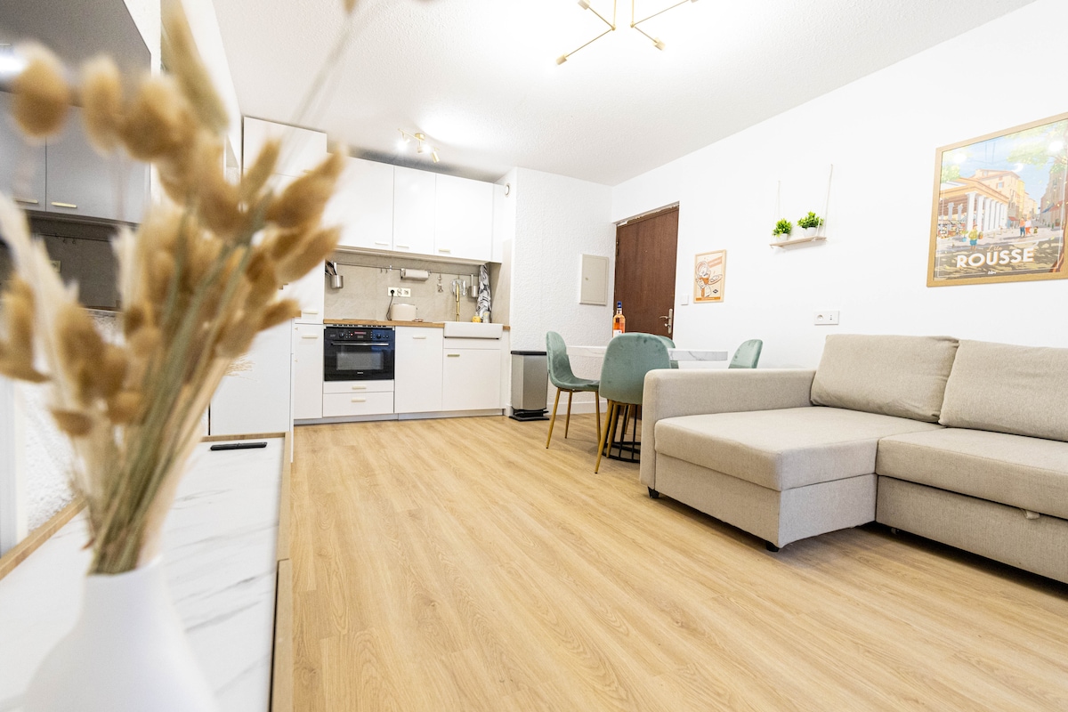 The open-plan living area features a modern kitchen equipped with essential appliances and a dining table with four chairs. A light-colored sectional sofa provides seating. Natural light fills the space through a window, creating an inviting ambiance.