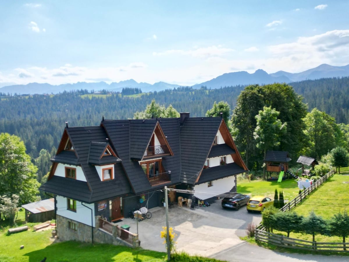 The exterior of a spacious multi-level house is showcased against a backdrop of rolling mountains and clear blue skies. The property features a well-maintained garden and parking area, with scenic greenery surrounding the building.