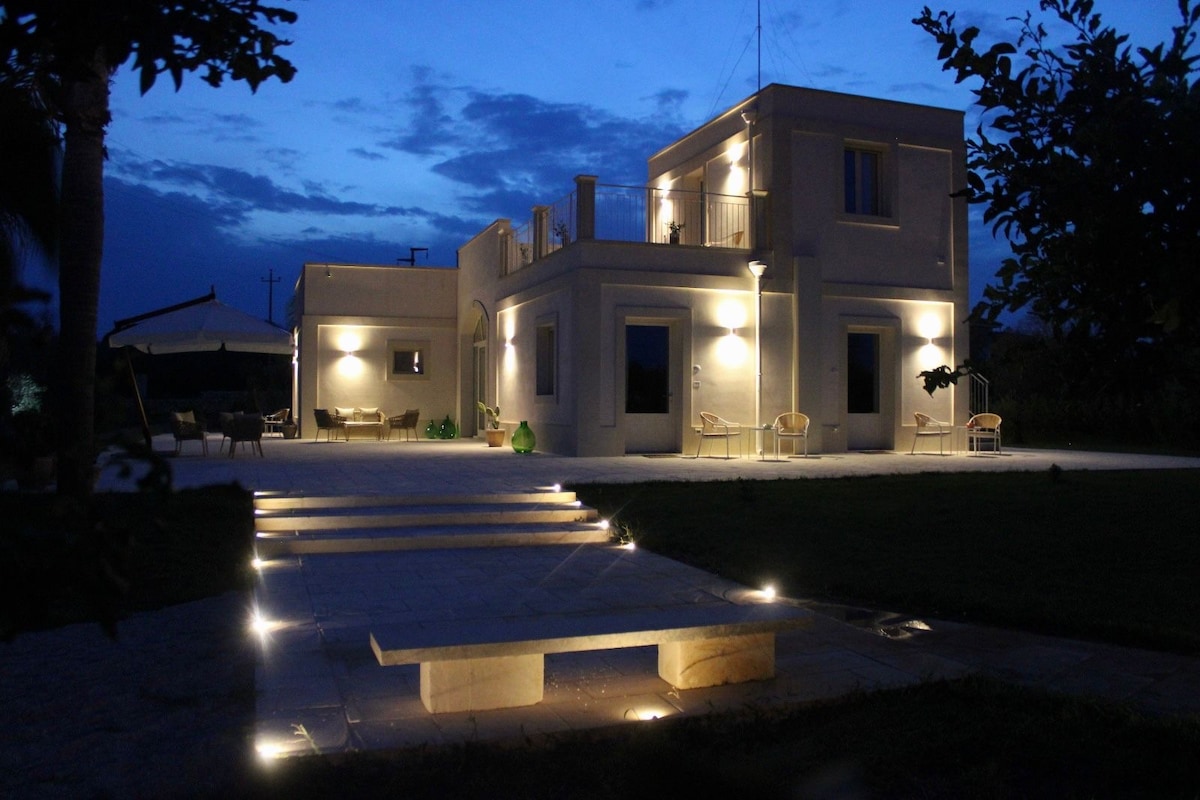 The elegant property is illuminated under a dusky sky, highlighting its classic architectural features. Soft lighting enhances the outdoor seating area and patio steps, creating a serene ambiance. Lush greenery surrounds the space, inviting relaxation and connection with nature.