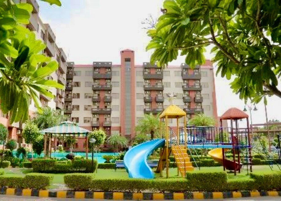 A well-maintained outdoor play area is depicted, featuring colorful playground equipment including slides and swings, surrounded by neatly trimmed greenery. A backdrop of a multi-story building is visible, providing a vibrant and welcoming environment for families.
