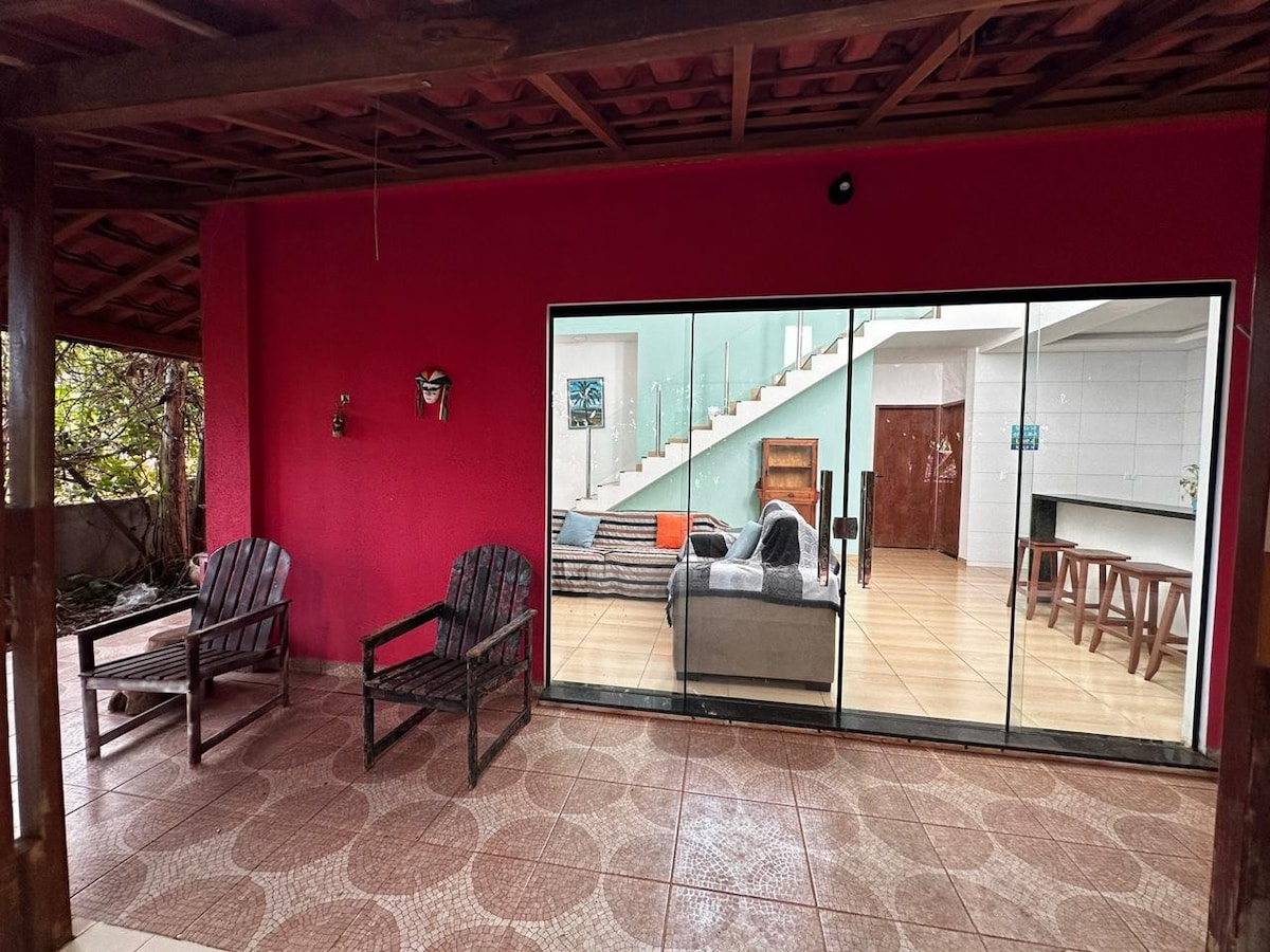 An outdoor seating area features two dark wooden chairs facing a large sliding glass door. The vibrant red wall complements the view of the interior, showcasing a staircase and a spacious living area filled with natural light.