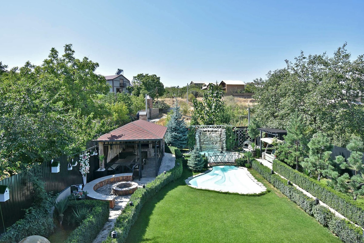 A spacious outdoor area is featured, showcasing a well-maintained lawn surrounded by lush greenery. A swimming pool is visible, accompanied by a covered seating area. A wooden gazebo is present, complemented by a stone pathway leading to various landscaped sections.