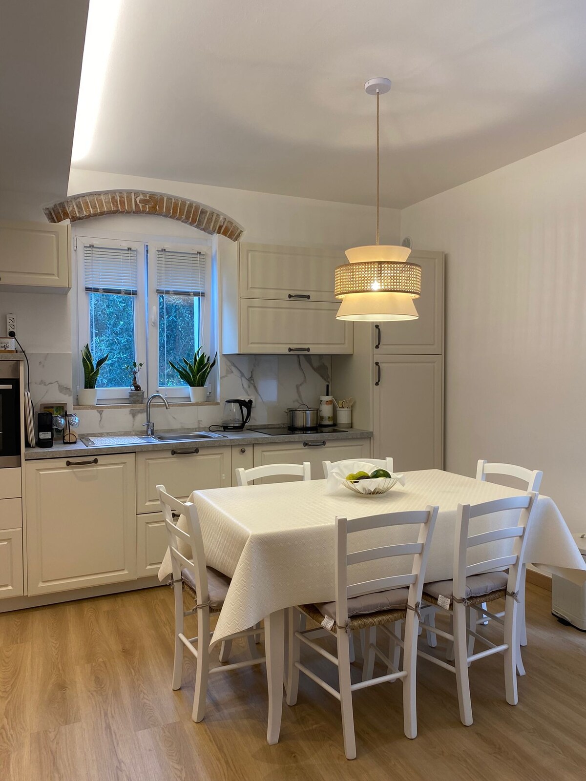 A clean and airy kitchen is seen, featuring a dining table surrounded by white chairs. Natural light enters through a window above the sink, complemented by plants. A light fixture hangs overhead, and cabinetry provides a modern and functional design.