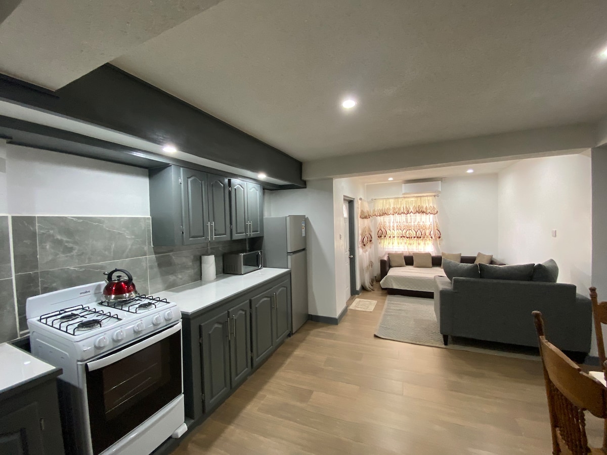 A modern kitchen area showcases dark cabinetry paired with white countertops. An oven and a microwave are visible, along with a red kettle on the stove. The open layout transitions to a cozy living space, featuring a gray sectional sofa and gentle lighting.