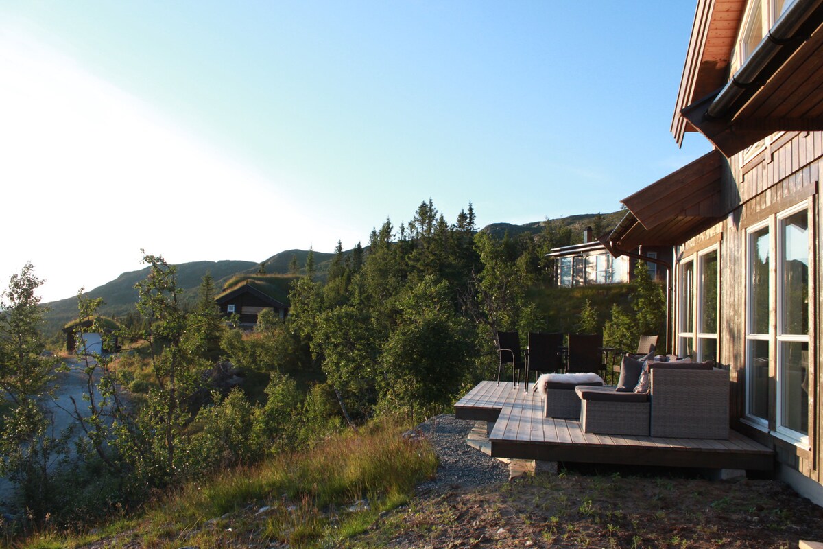 A spacious terrace is presented, featuring comfortable outdoor seating arrangements. The surrounding landscape showcases lush greenery and distant hills, allowing for scenic views. The gentle sunlight highlights the wooden structure, creating a welcoming outdoor space for relaxation.