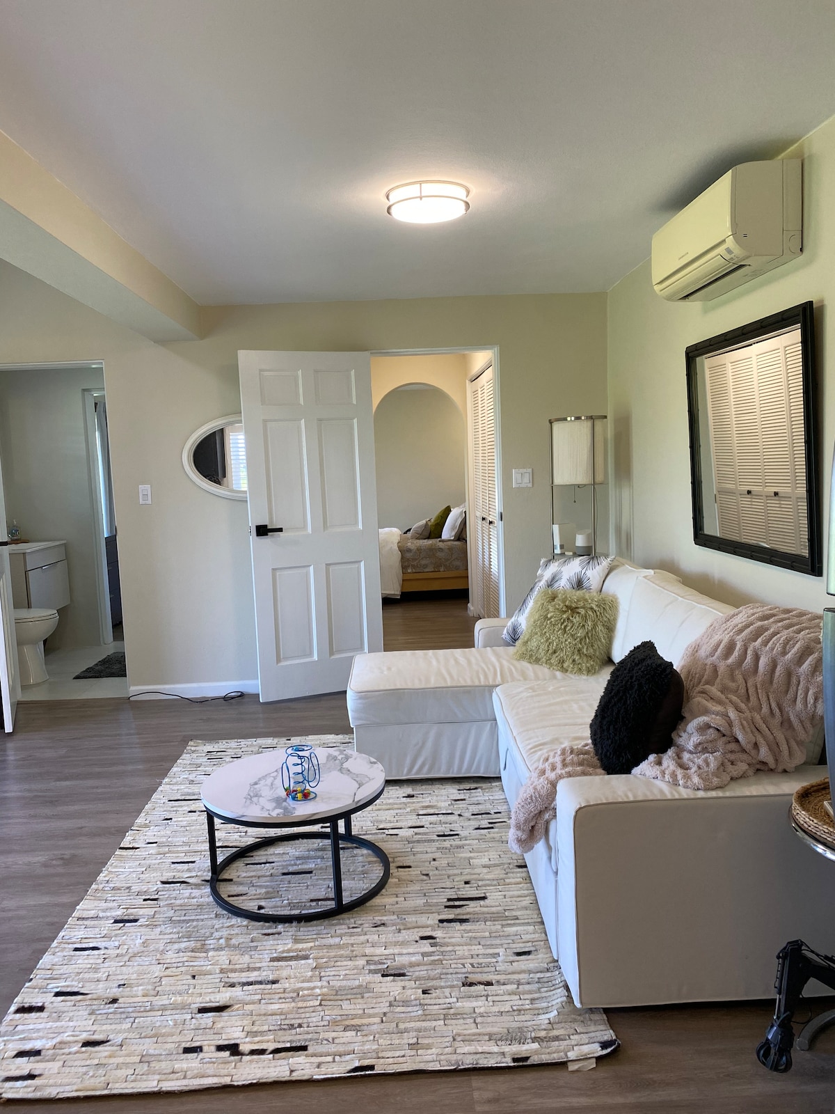 A light-filled living area is seen with a white sofa adorned with textured pillows. A round coffee table sits on a woven rug, while the backdrop reveals a hallway leading to a cozy sleeping area featuring a framed mirror and neutral tones.