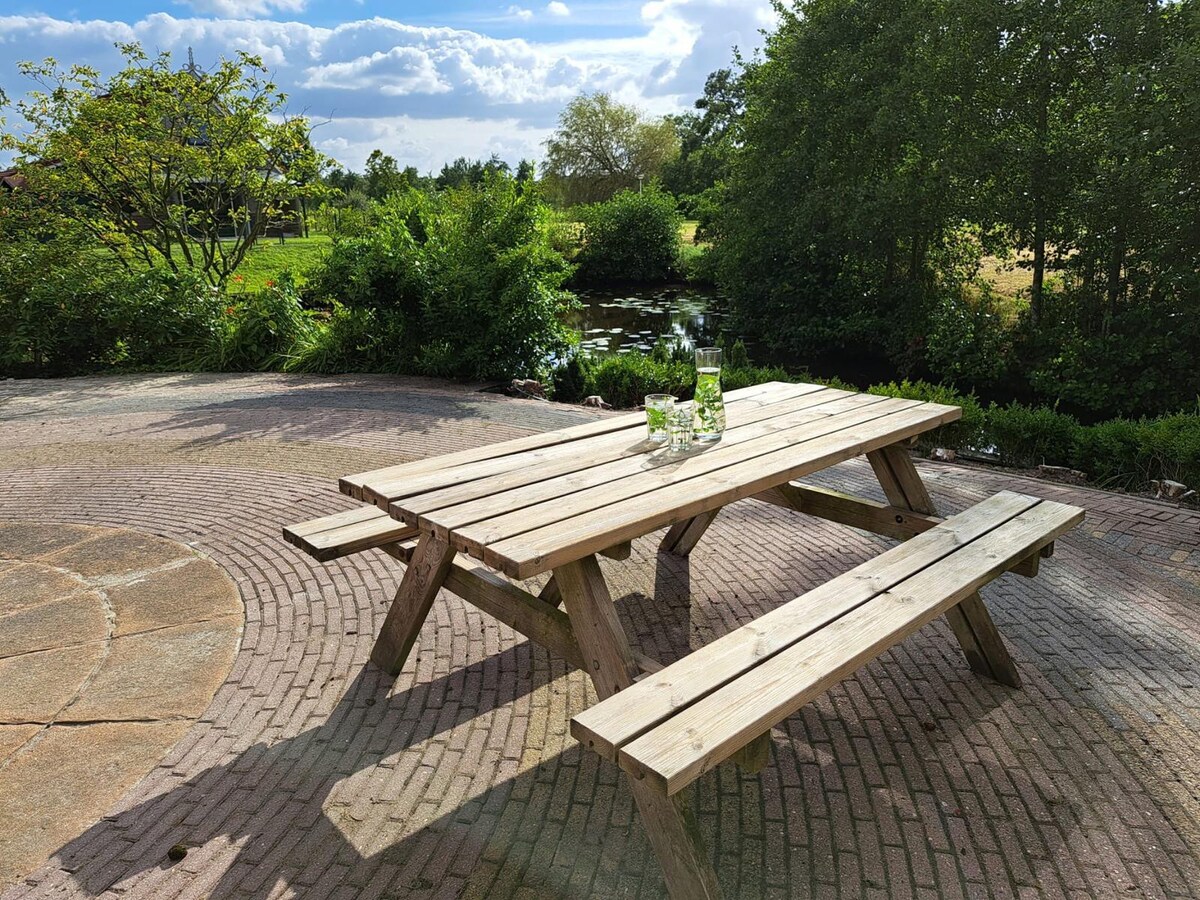 A wooden picnic table with attached benches is positioned on a paved area. Glass bottles sit on the table, with lush greenery and a pond visible in the background. The setting is illuminated by soft natural light, creating a serene outdoor space.