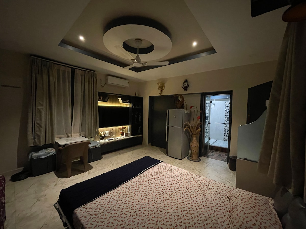 A cozy bedroom is highlighted by a spacious bed adorned with a patterned cover. Soft lighting creates a warm atmosphere, while a functional workspace with a desk and chair is visible. A refrigerator stands nearby, and a bathroom entryway can be seen in the background.