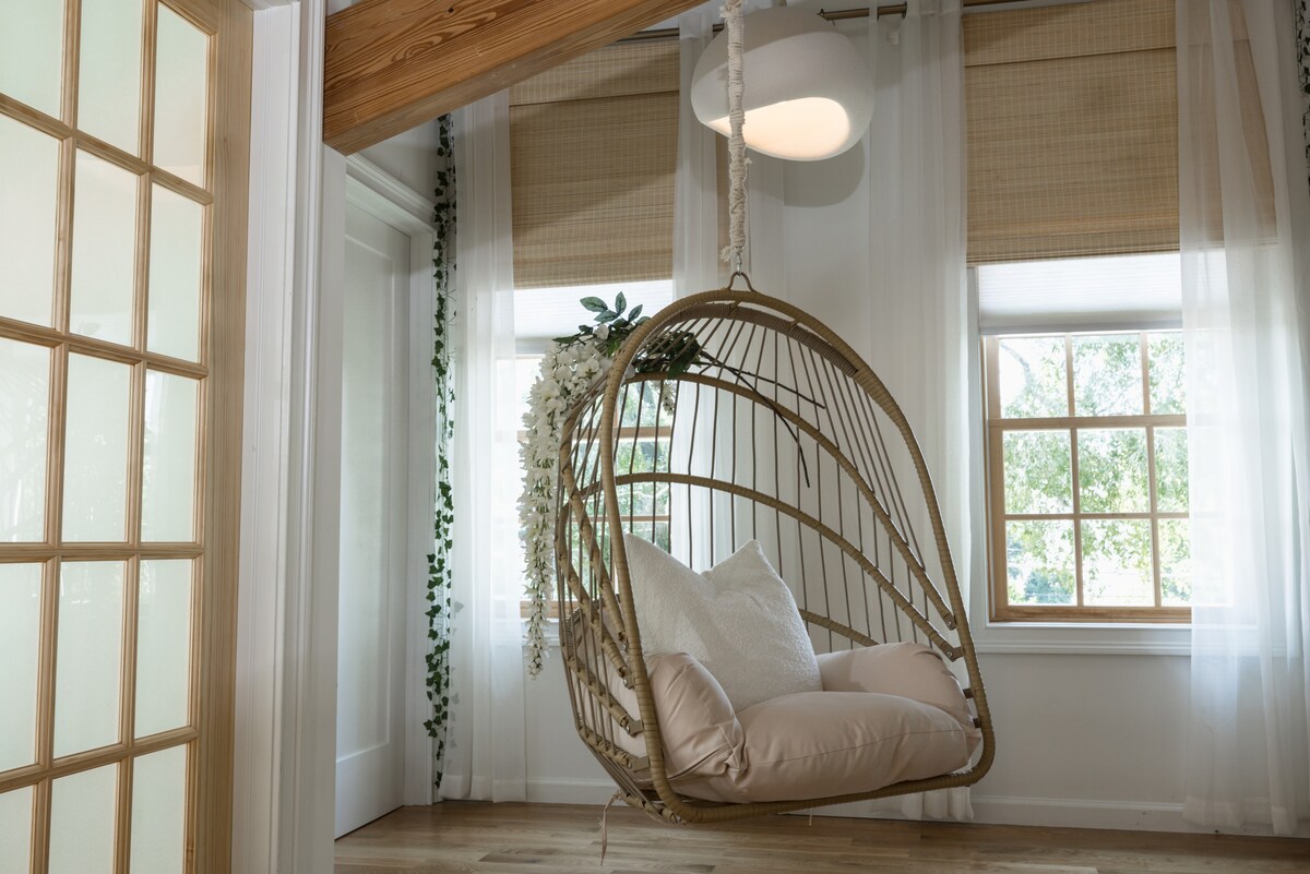 A suspended swing chair is featured in this airy space, surrounded by natural light filtering through large windows. Soft pink cushions provide comfort, while leafy accents are visible in the background. The room is highlighted by neutral tones and inviting wooden beams.