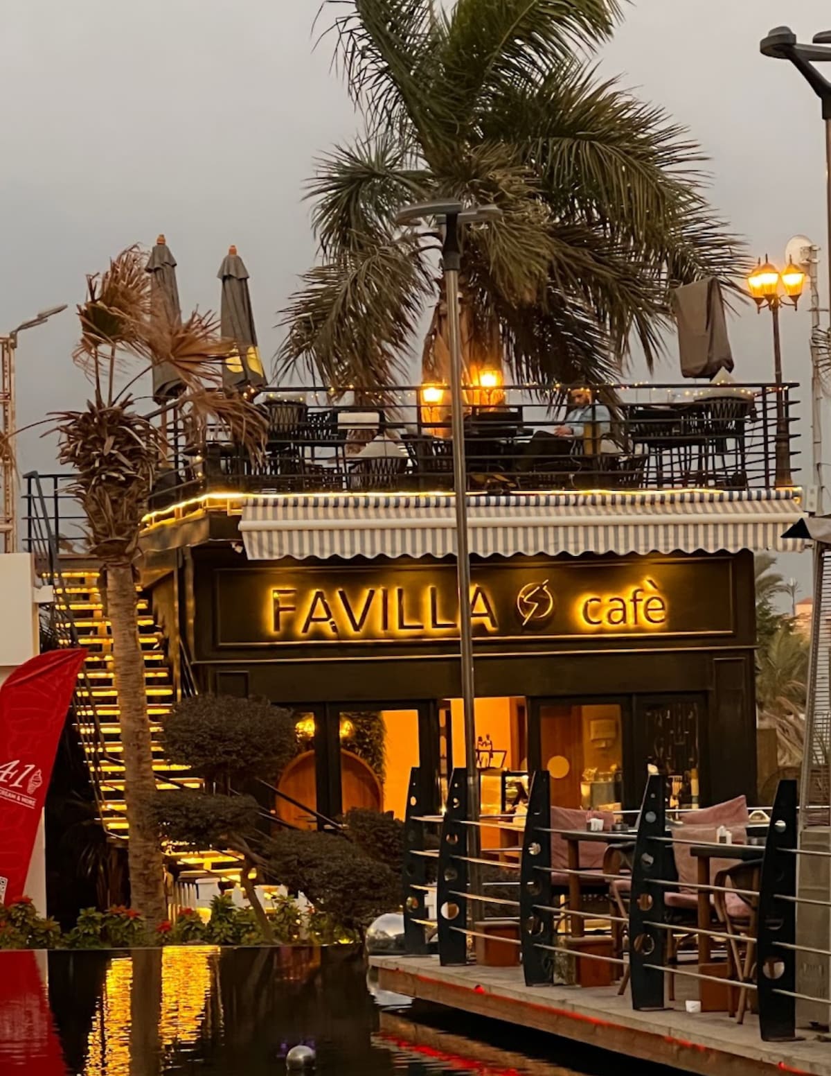 A two-story café building features a modern exterior with large windows and a warm glow from the interior lights. Outdoor seating is arranged on the upper terrace, shaded by market umbrellas, surrounded by palm trees and decorative lighting.