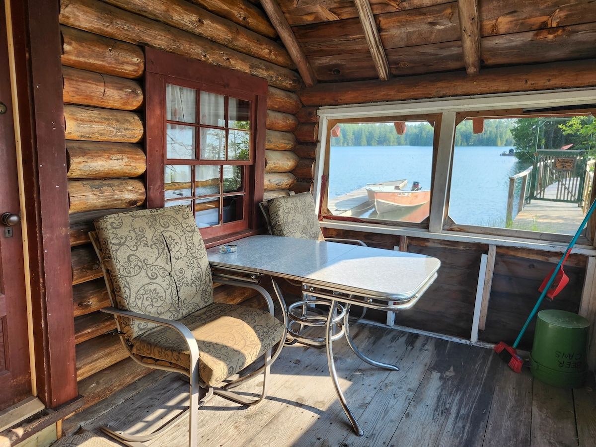 A screened porch features two cushioned chairs and a dining table, providing a space for relaxation with views of the lake. The log cabin walls add a rustic touch, while the large windows offer natural light and a serene backdrop of water and boats.