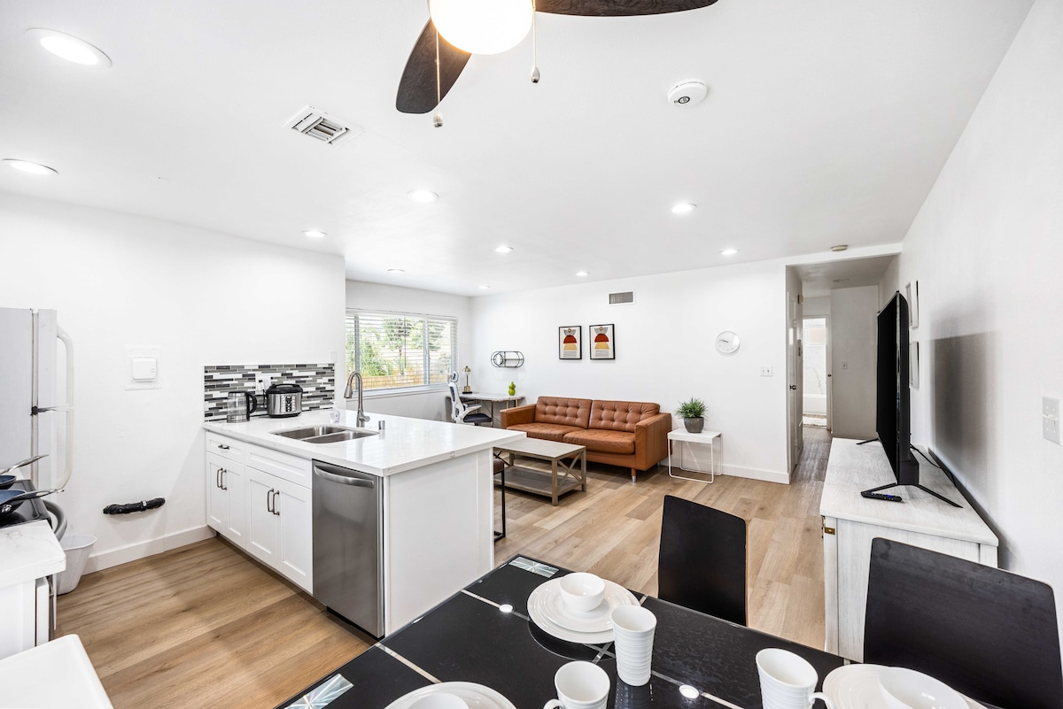 An open living space is shown, featuring a modern kitchen with sleek white cabinetry and a spacious island. A comfortable brown sofa complements the seating area, while a dining set with tableware is arranged for four. Natural light fills the room through a large window.