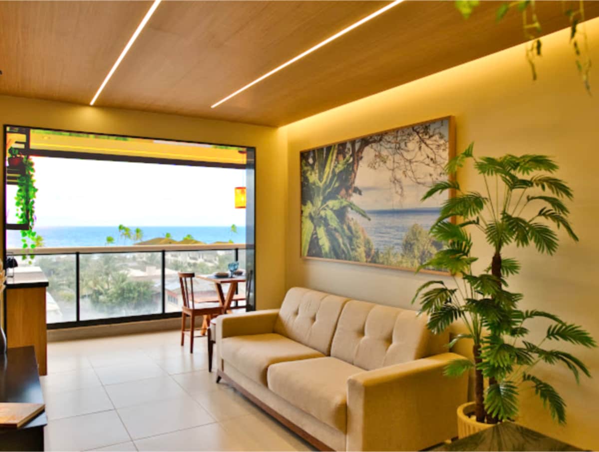 A modern living area features a beige sofa positioned near large glass doors that open to a stunning sea view. Natural light fills the space, highlighting a tropical-themed artwork on the wall and a small dining set nearby. Greenery adds a refreshing touch.