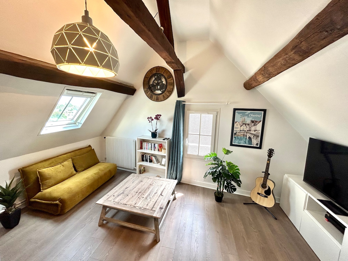 The living area features a high, sloped ceiling with exposed wooden beams. A bright yellow sofa faces a wooden coffee table, complemented by a small plant and an acoustic guitar nearby. Natural light is allowed in through a window dressed with light curtains.
