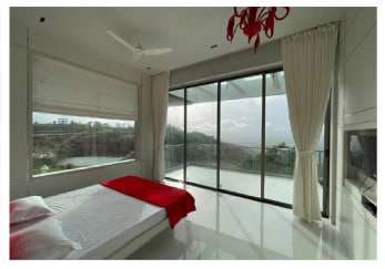 A bright bedroom features a spacious bed dressed in white linens, complemented by a bold red throw. Large glass doors invite natural light and provide views of the surrounding landscape, while sheer curtains frame the window, enhancing the open and airy feel of the space.