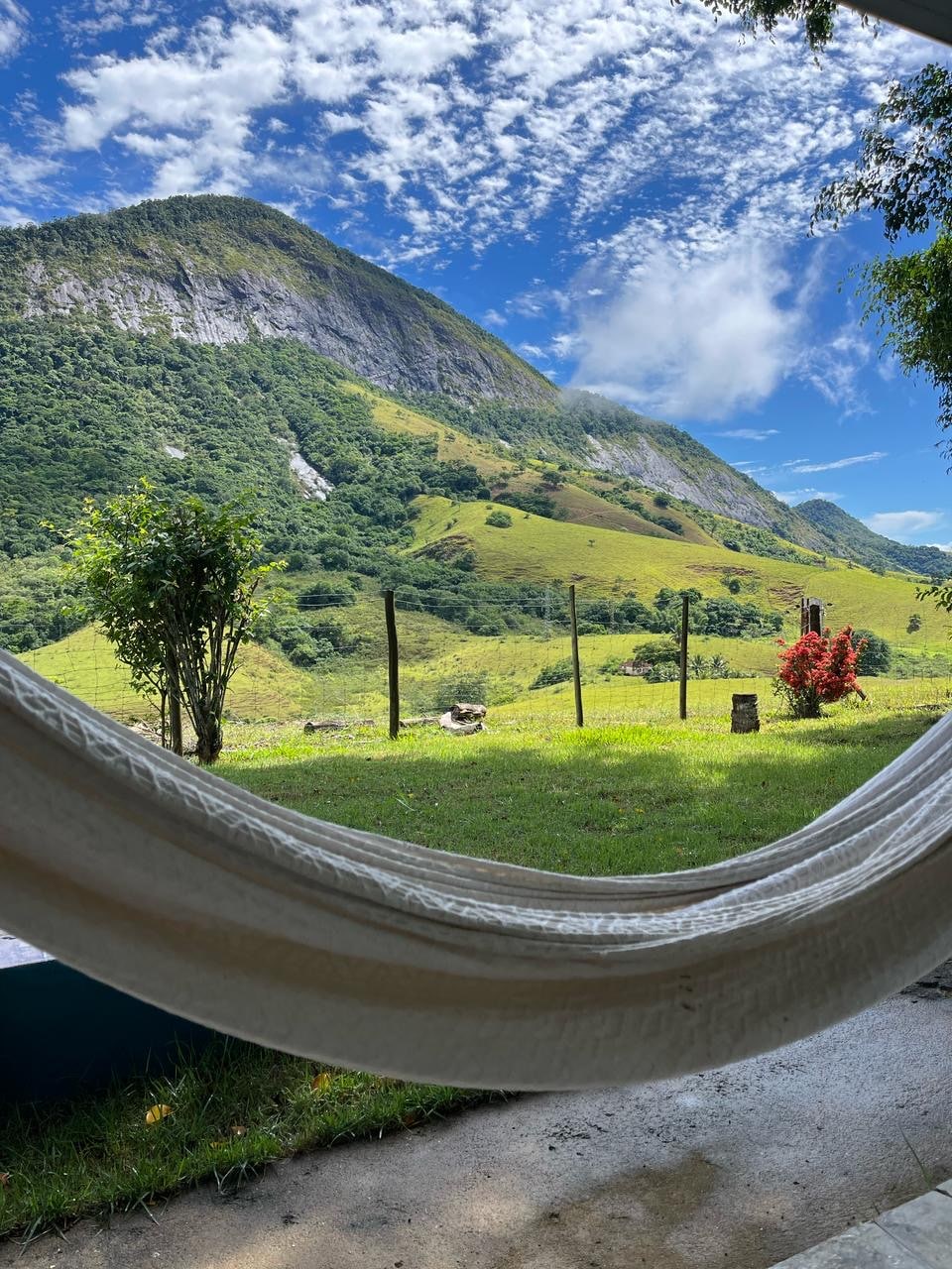 A tranquil view from a hammock captures the surrounding mountains and lush greenery. The vibrant colors of flowers are visible in the foreground, complemented by a clear blue sky dotted with clouds. The landscape showcases the natural beauty of the area.