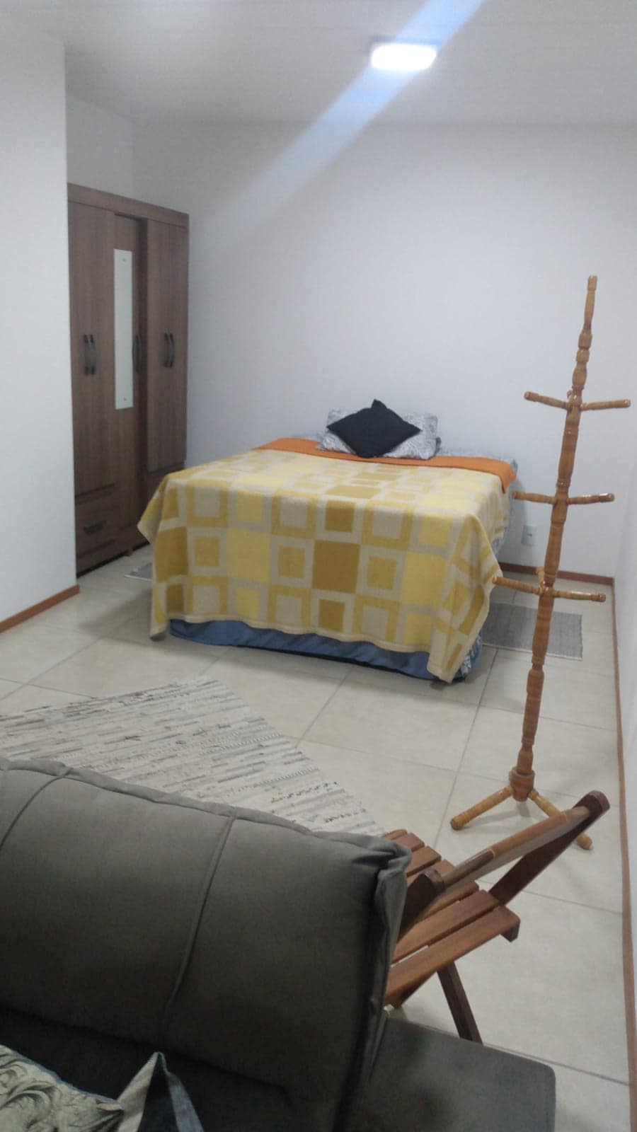 A cozy room features a bed covered with a yellow blanket and a decorative black pillow. A wooden wardrobe is positioned nearby, and a light-toned rug lies on the tiled floor. A folding chair and a coat rack are present, contributing to the room's functionality.