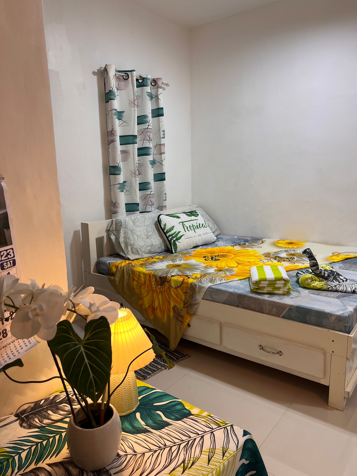 A cozy sleeping area features a light-colored bed adorned with a vibrant floral blanket. A decorative pillow with the word 'Tropical' rests on the bed. A small table with a plant and a lamp is positioned beside the bed, complementing the bright, cheerful decor.