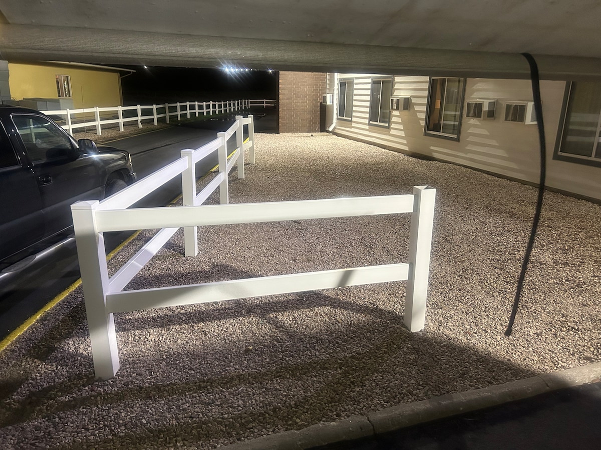 A well-maintained gravel parking area is bordered by a white wooden fence. A section of the building is visible in the background, with several air conditioning units positioned on the exterior wall.