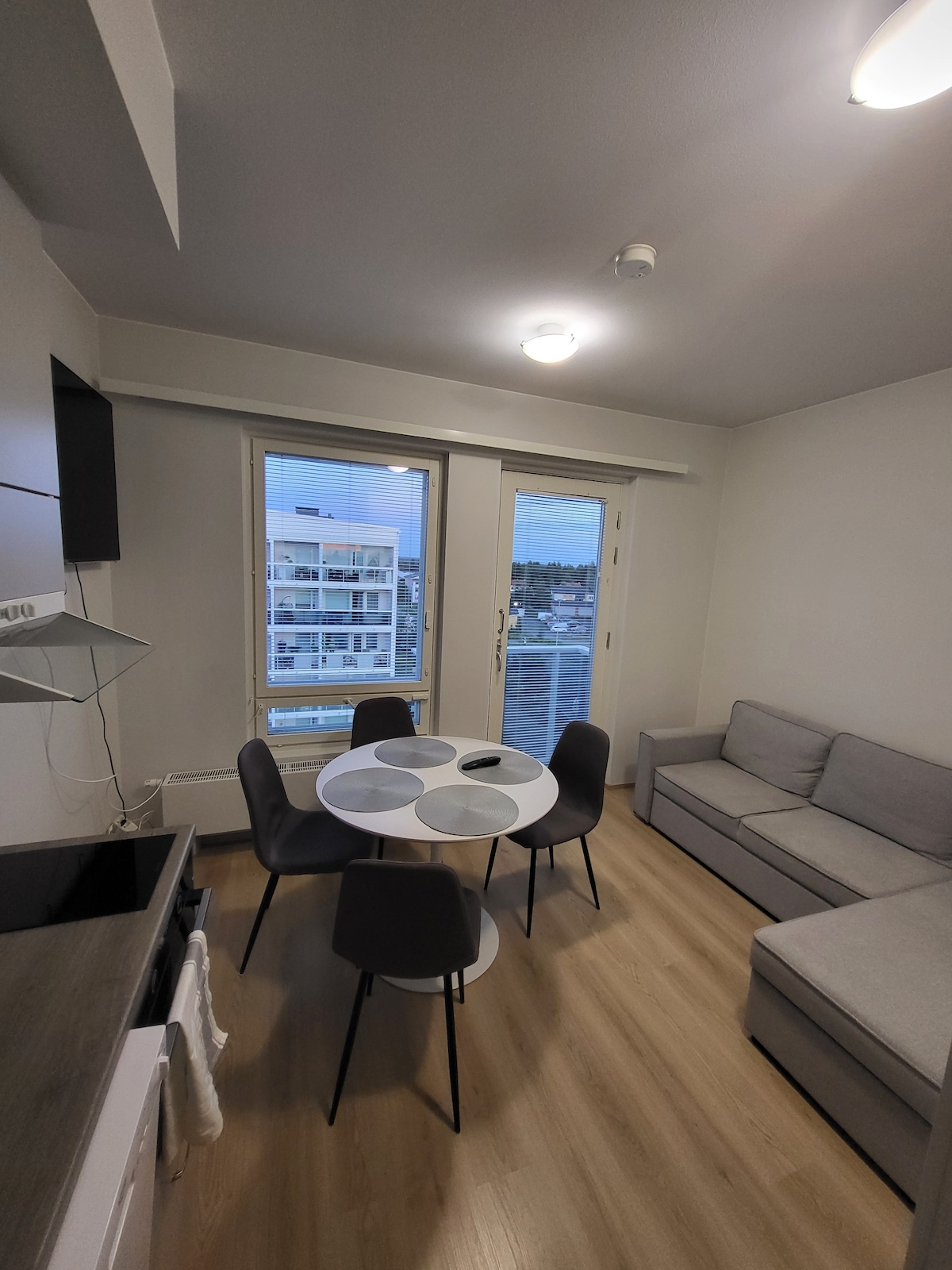 An open-plan living area is featured, showcasing a light-colored sofa and a round dining table surrounded by four chairs. Large windows provide a view of the outside, allowing natural light to fill the space. The kitchen area is partially visible with modern appliances.