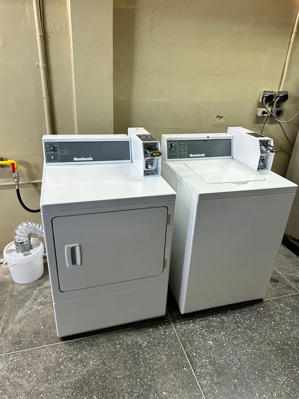 A laundry area features a stacked dryer and washer, both in a light grey finish. The appliances are positioned against a neutral wall, with a small plant in a white pot nearby. The floor is covered in dark, speckled tiles.