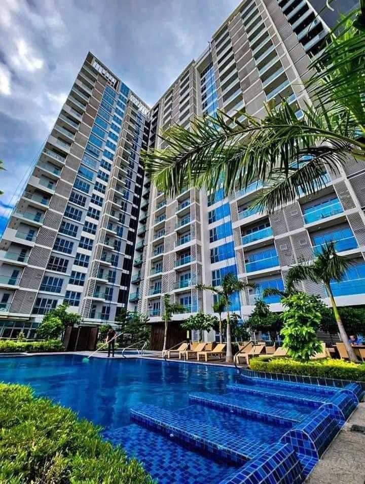 A modern residential building is depicted with multiple stories and large glass windows, surrounded by lush greenery. A blue swimming pool with steps leading in creates a serene outdoor space, complemented by lounge chairs under palm trees.