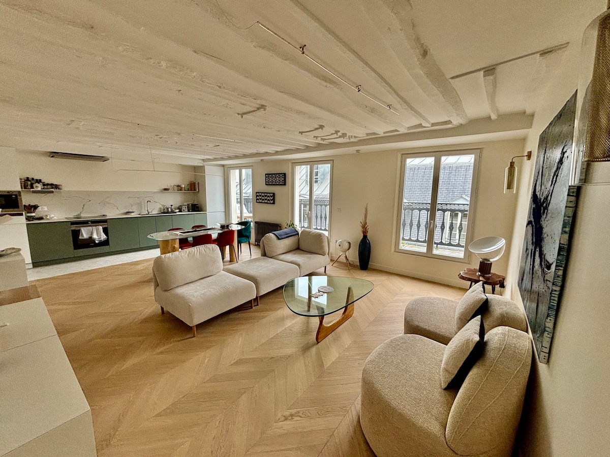The living area features a combination of modern furniture design, including soft, neutral-toned seating arranged around a glass coffee table. Natural light floods the space through multiple windows, highlighting the light wood flooring and sleek kitchen layout in the background.