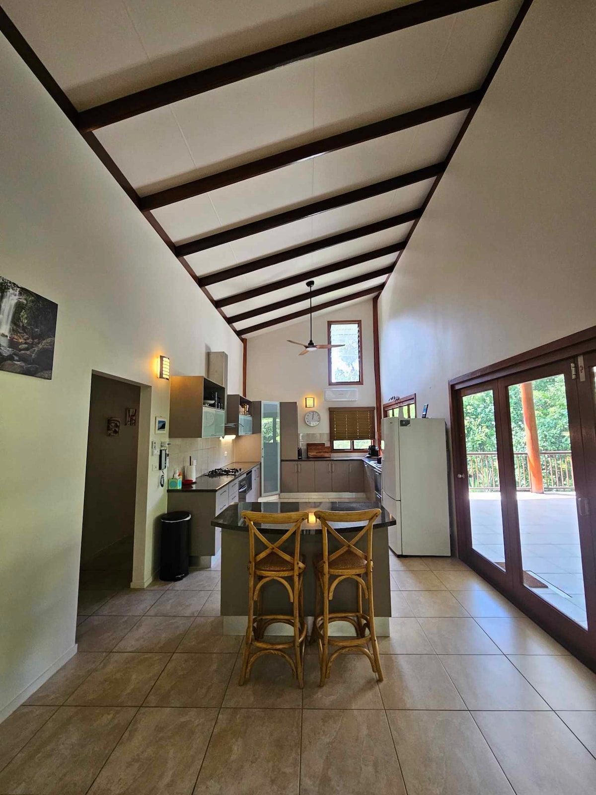 The spacious kitchen features high ceilings with exposed beams and an island countertop with seating. Natural light fills the area through large windows, which offer views of the surrounding greenery. Modern appliances and cabinetry create a functional and inviting space for meal preparation.