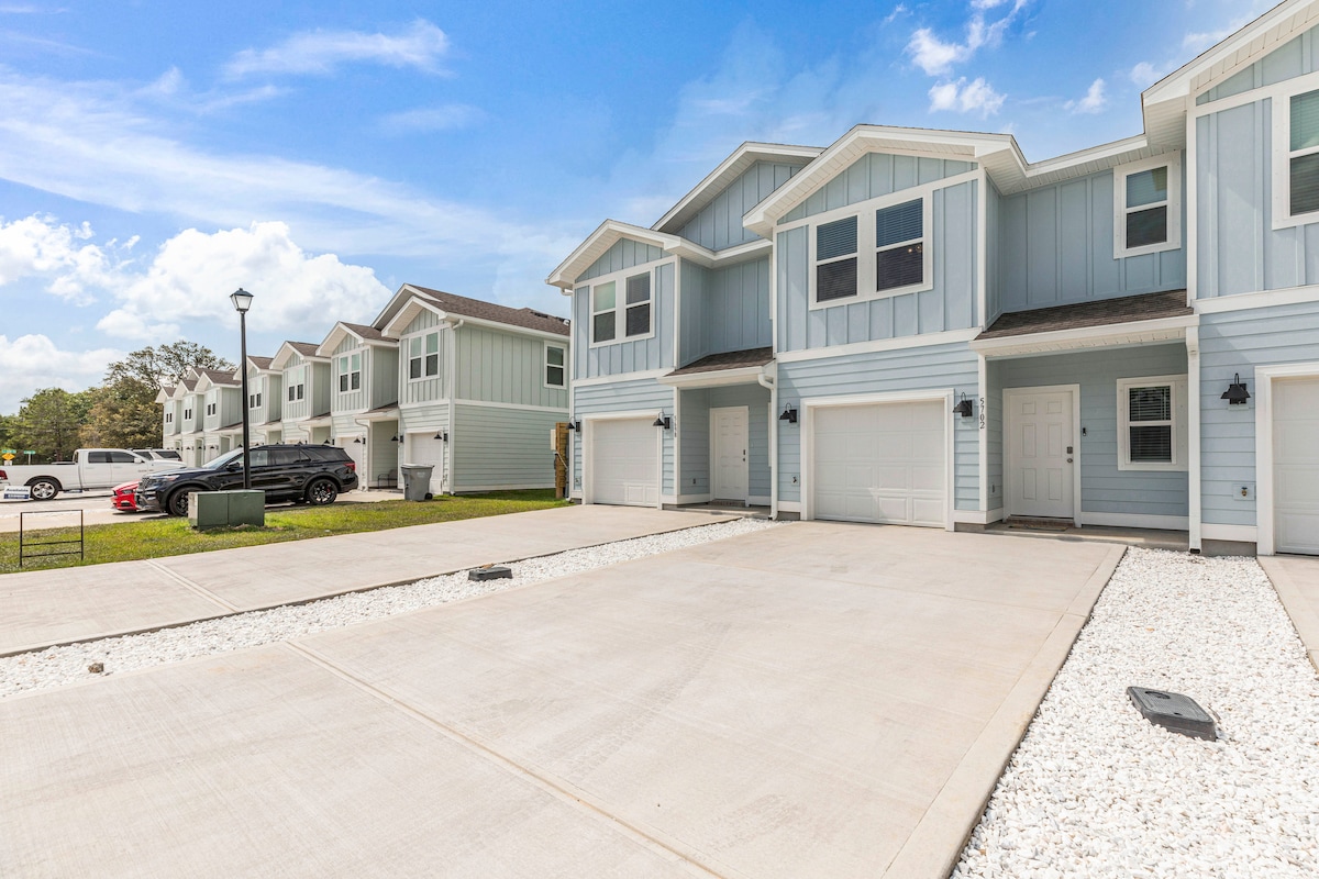 A newly constructed townhome exterior is showcased, featuring a light blue façade and a spacious driveway. Multiple townhomes are visible in a well-maintained community setting under a clear blue sky. A grassy area is located nearby, enhancing the neighborhood's appeal.