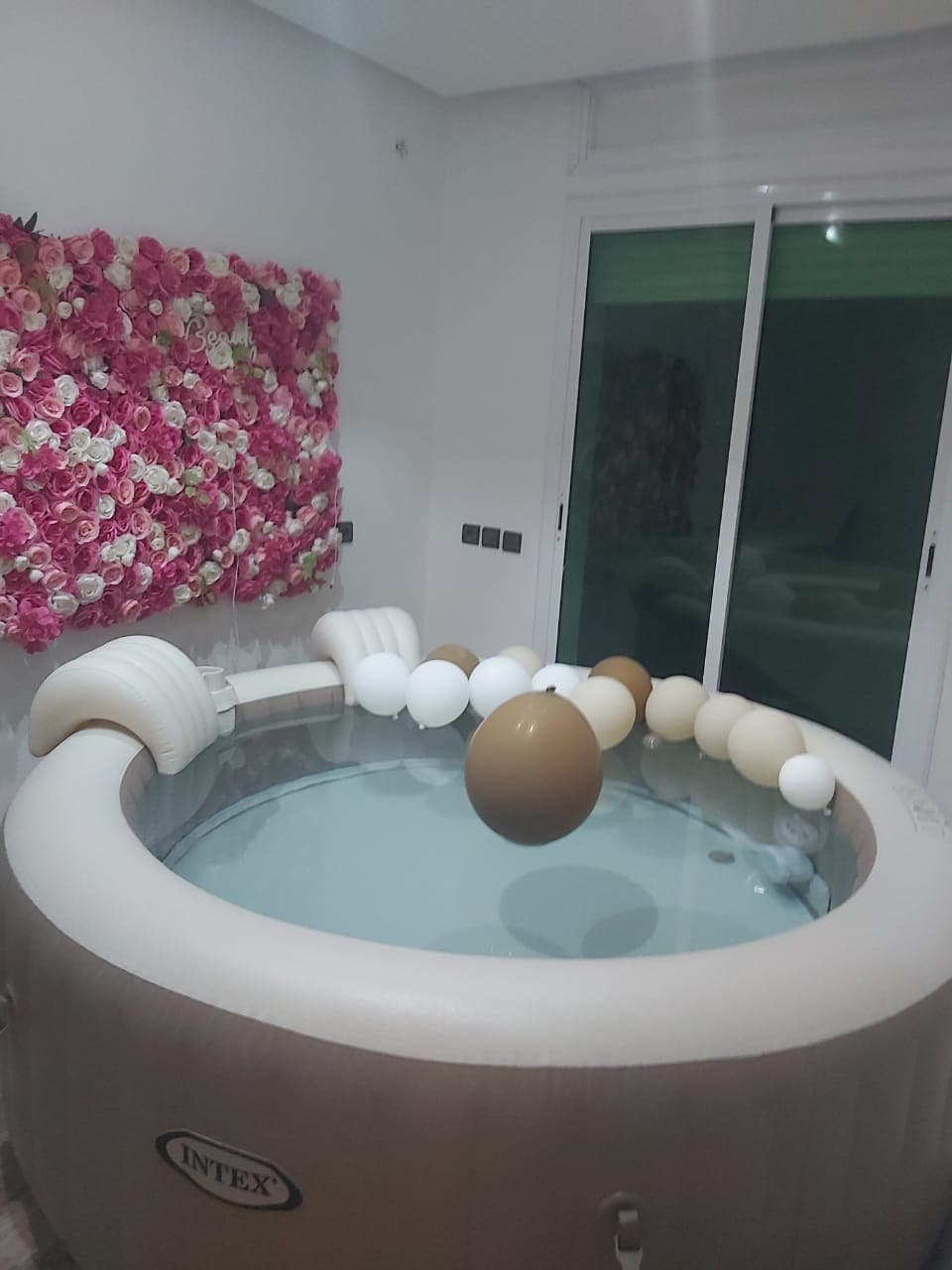 A circular inflatable jacuzzi is positioned indoors, surrounded by decorative balloons in neutral tones. A vibrant flower wall with pink and white blooms is displayed in the background, while large glass doors provide access to the outside, allowing natural light to enter the space.