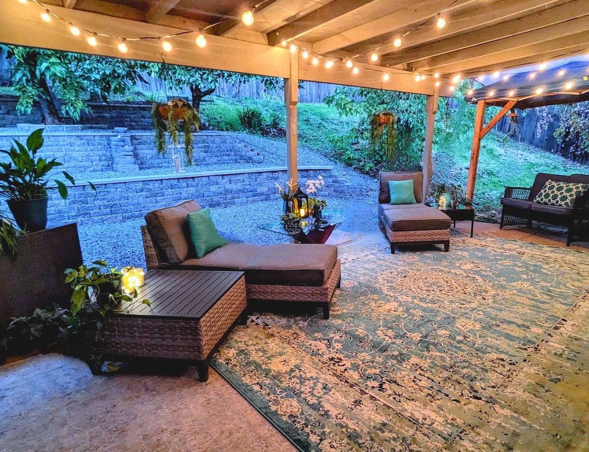 A covered patio area features two lounge chairs adorned with green cushions. A decorative rug adds warmth to the space. String lights hang above, creating a soft ambience. Potted plants contribute to the serene atmosphere, and a landscaped area with steps is visible in the background.