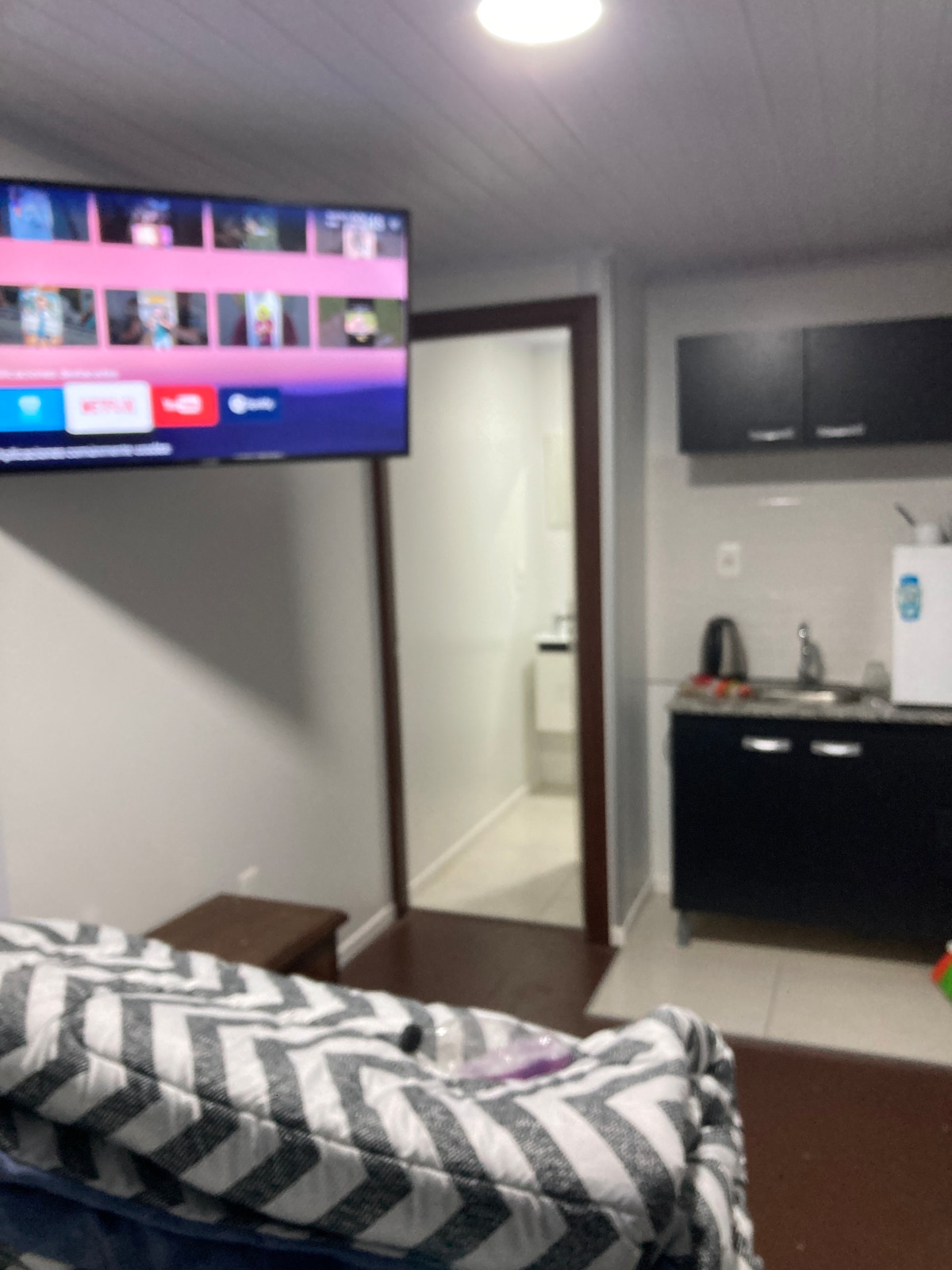 A compact living area is shown, featuring a wall-mounted television displaying various streaming options. A kitchenette with a sink and small appliances is adjacent, complemented by dark cabinetry. A bathroom door is visible in the background, providing easy access.