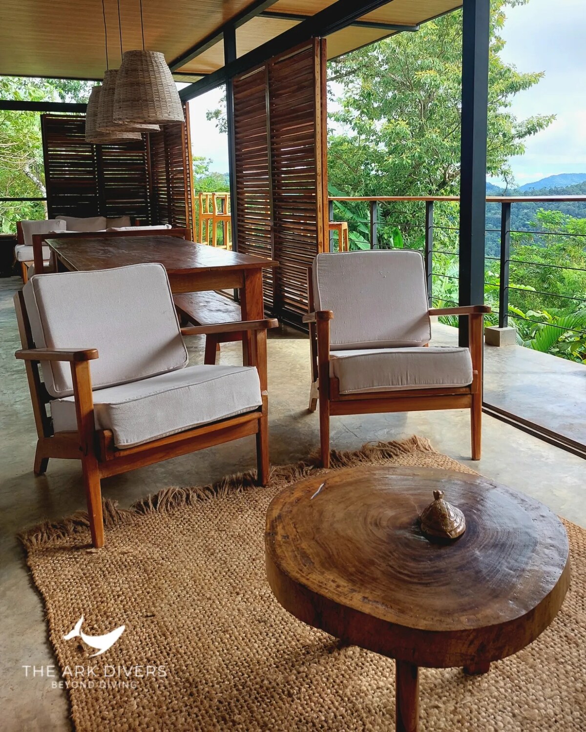 Two wooden chairs with light gray cushions are positioned beside a rustic wooden coffee table. A natural fiber rug lies underneath, and large glass doors offer views of the surrounding greenery, letting in ample natural light.