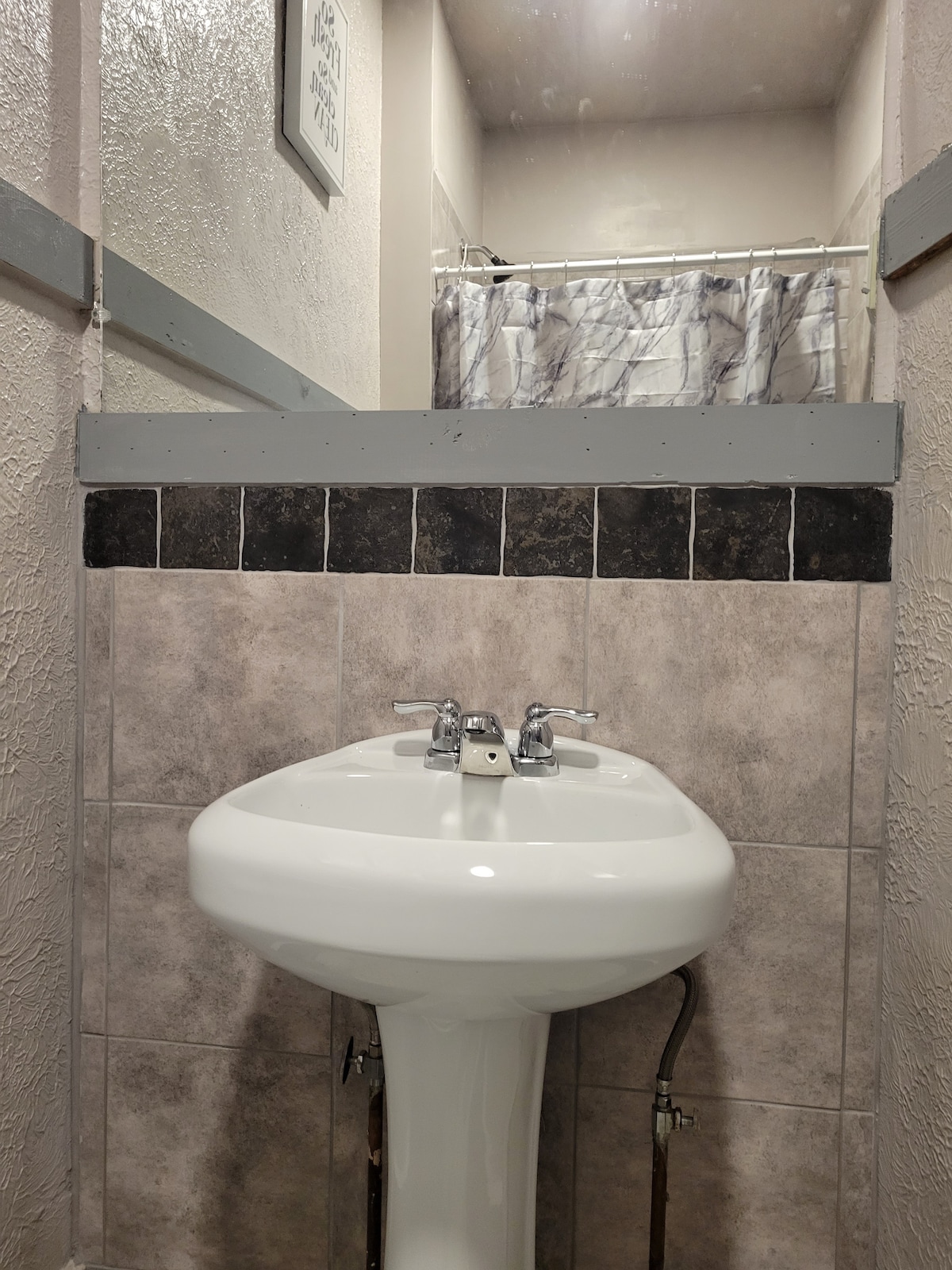 A compact bathroom area features a white pedestal sink with dual faucets. The walls are adorned with light gray and patterned tiles. A shower area is visible in the background, enclosed by a curtain with a subtle design.