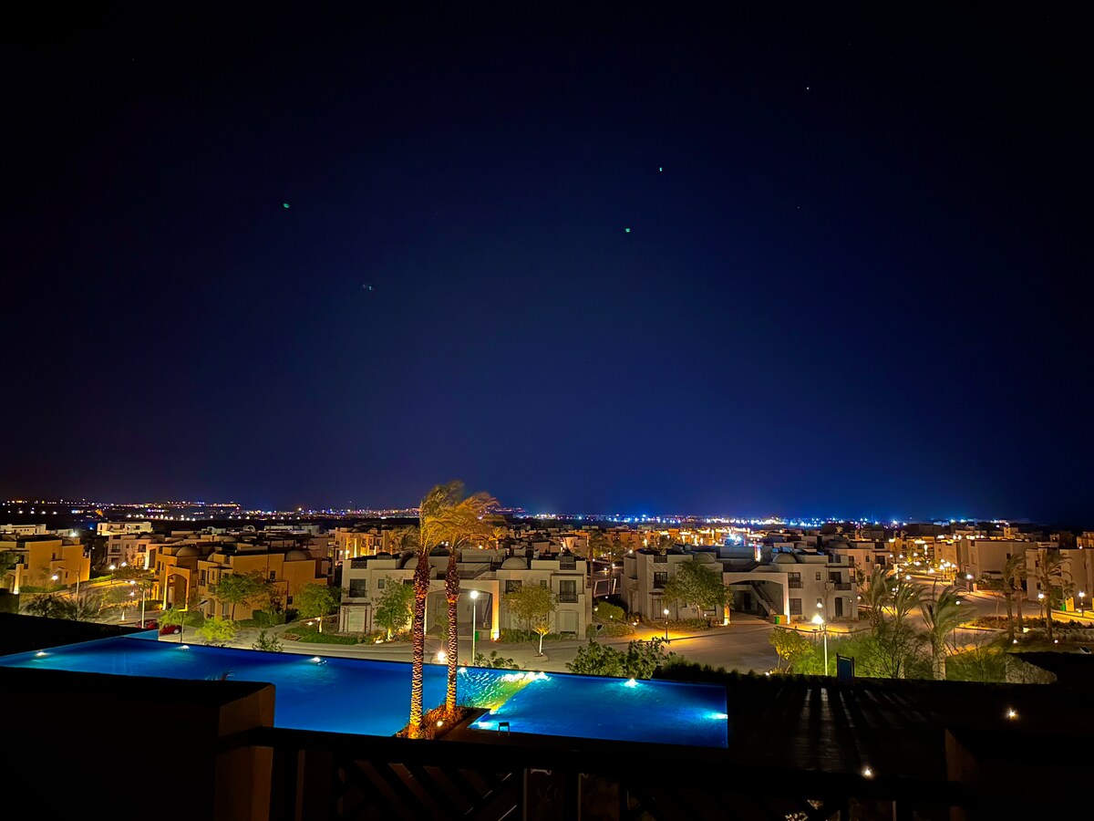 An expansive night view captures illuminated homes nestled in sprawling landscapes, with warm lights twinkling against a dark sky. A serene pool appears in the foreground, reflecting the city lights and adding tranquility to the vibrant scene.