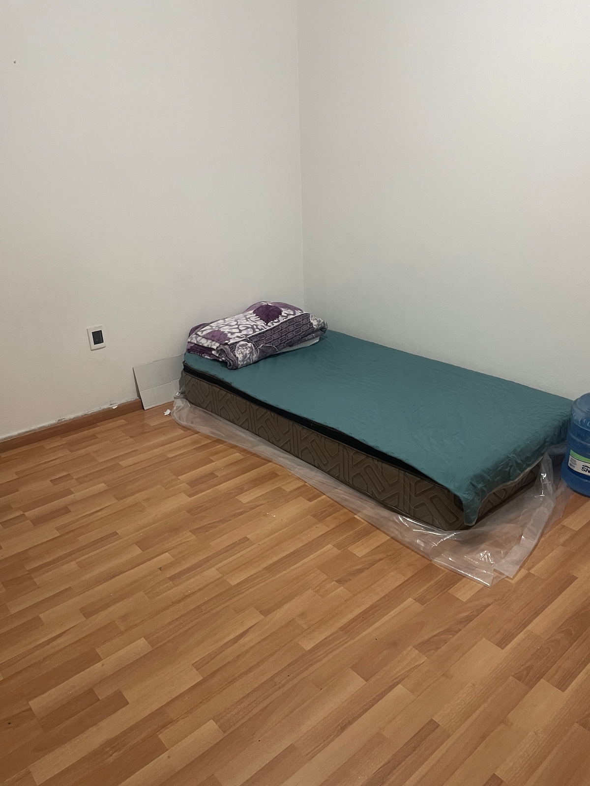 A simple sleeping space is presented, featuring a mattress placed on a wooden floor, covered with a green mattress protector. A folded blanket with a decorative pattern rests at the head of the bed. A water cooler is positioned next to the bed, providing convenience.