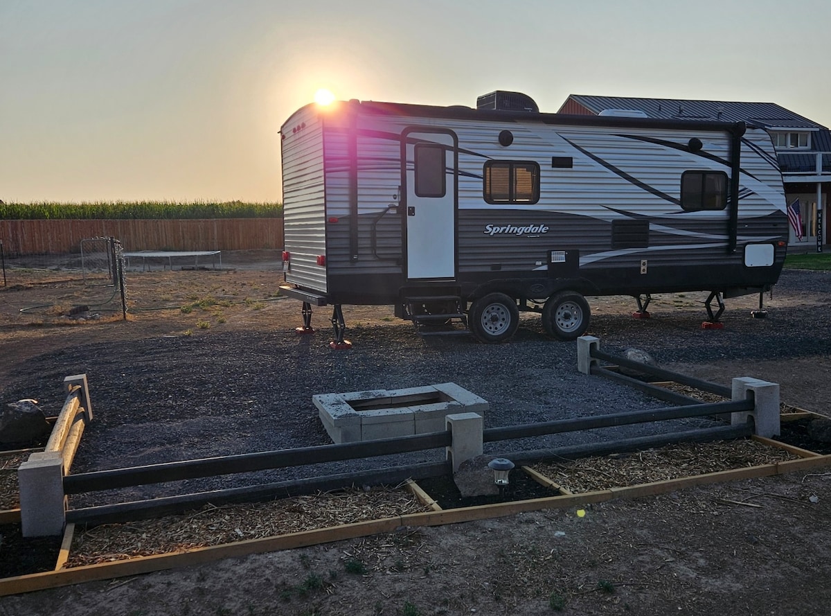 A travel trailer is positioned on a gravel surface, showcasing its sleek design with gray and black accents. The sun sets in the background, casting warm light, while a fire pit constructed from stone blocks is visible nearby, surrounded by a wooden frame.