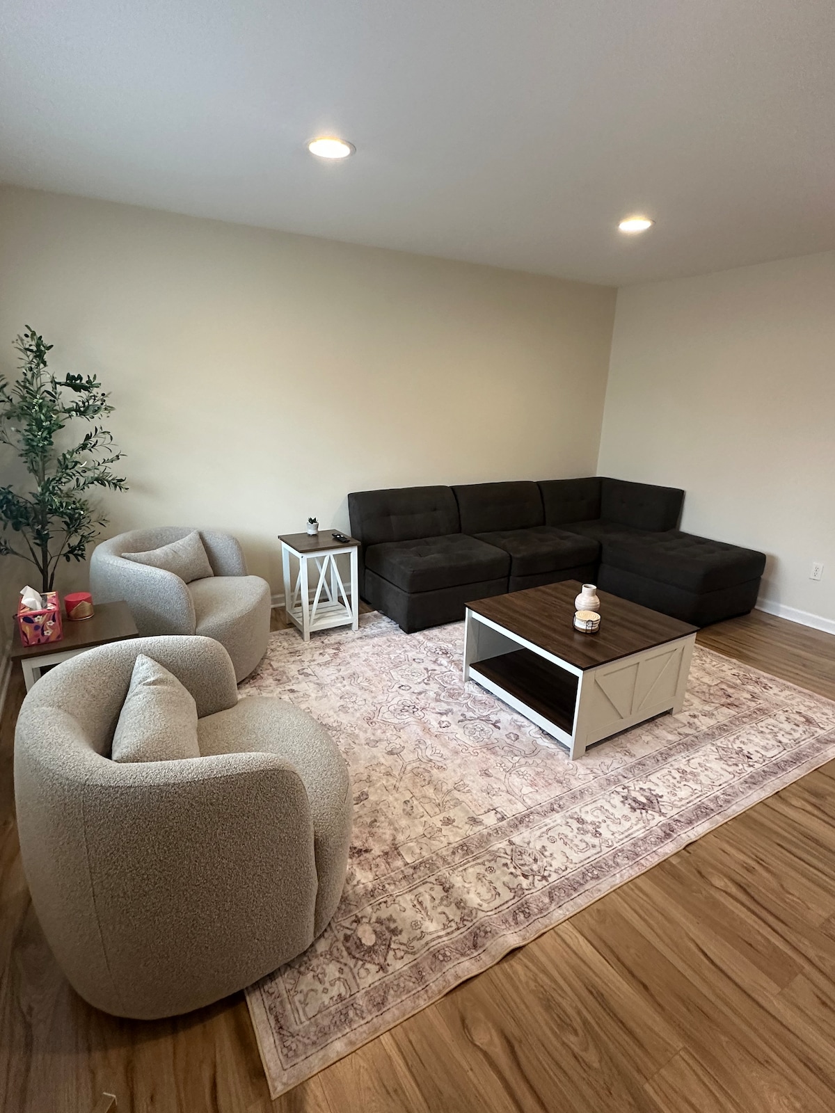 A welcoming living room features a dark sectional sofa alongside a light-colored coffee table. Plush, rounded armchairs provide additional seating. A soft area rug with a delicate pattern is layered over warm wood flooring, complemented by a small side table and a decorative plant.