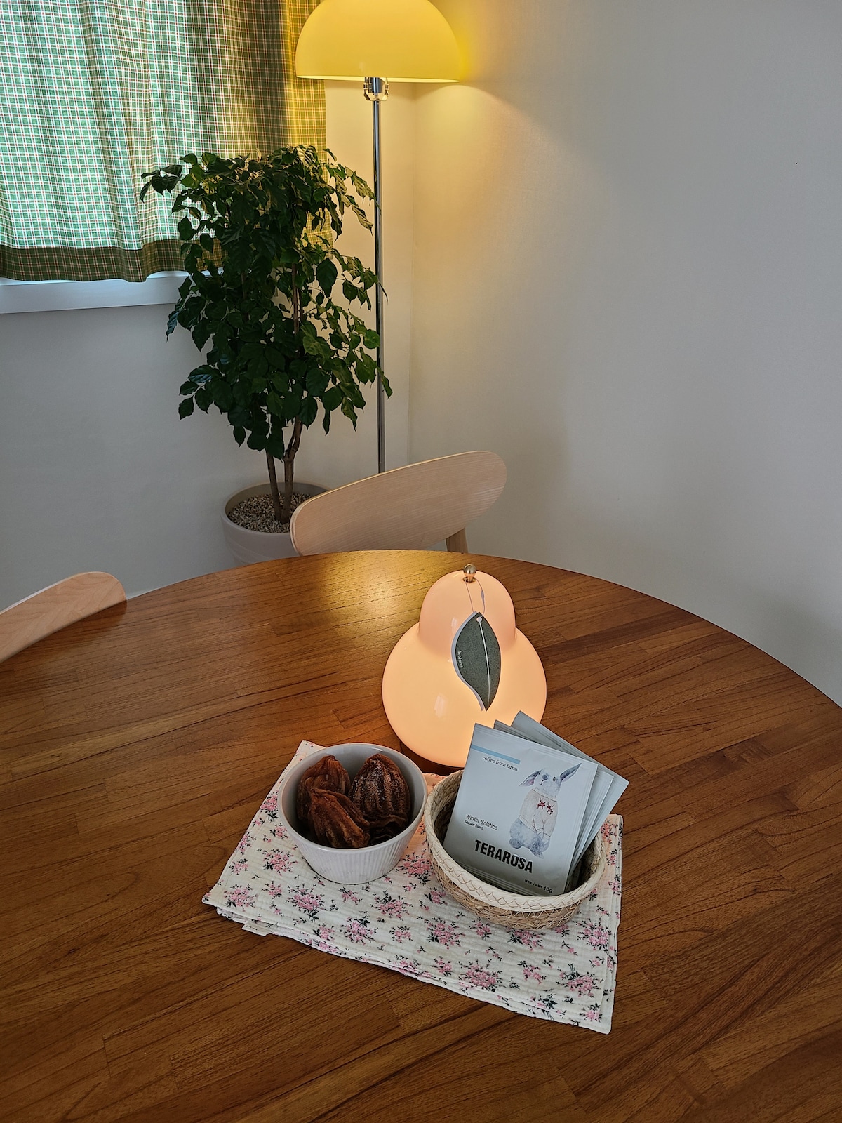 A round wooden dining table is set with a floral patterned tablecloth. Two decorative bowls are filled with pastries, and a booklet sits neatly beside them. A pear-shaped lamp casts a soft light, while a potted plant and green-checkered curtains are visible in the background.
