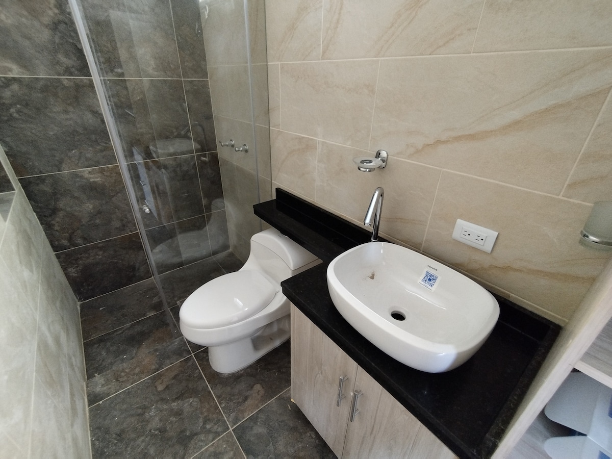 A modern bathroom is displayed, featuring a walk-in shower with clear glass walls and a sleek black stone countertop. A round sink is noted, accompanied by a flush toilet. Neutral-toned tiles cover the walls and floor, creating a clean and spacious appearance.