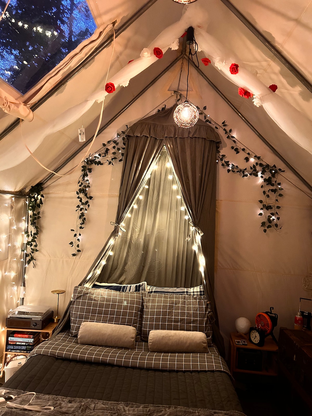 An elegantly furnished tent interior features a spacious bed adorned with checkered bedding and two accent pillows. Soft string lights and decorative elements, including greenery and floral accents, create a warm ambiance. A window allows natural light to filter through, enhancing the cozy atmosphere.
