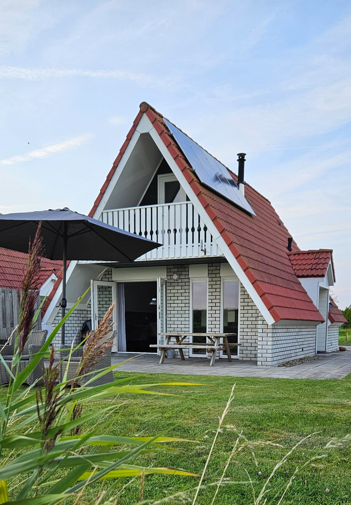 A charming two-story house with a distinctive triangular roof showcases large glass doors that open to a spacious patio. Natural elements like grass and tall plants frame the property, while a shaded outdoor table and umbrella provide a comfortable spot for relaxation.
